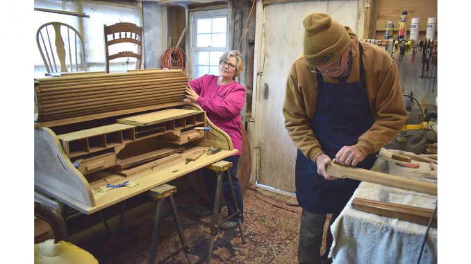 Susan Price at work in the Furniture Magicians workshop, steadying the dismantled carcase of a roll-top desk while a technician shapes a length of trim at the bench beside her.
