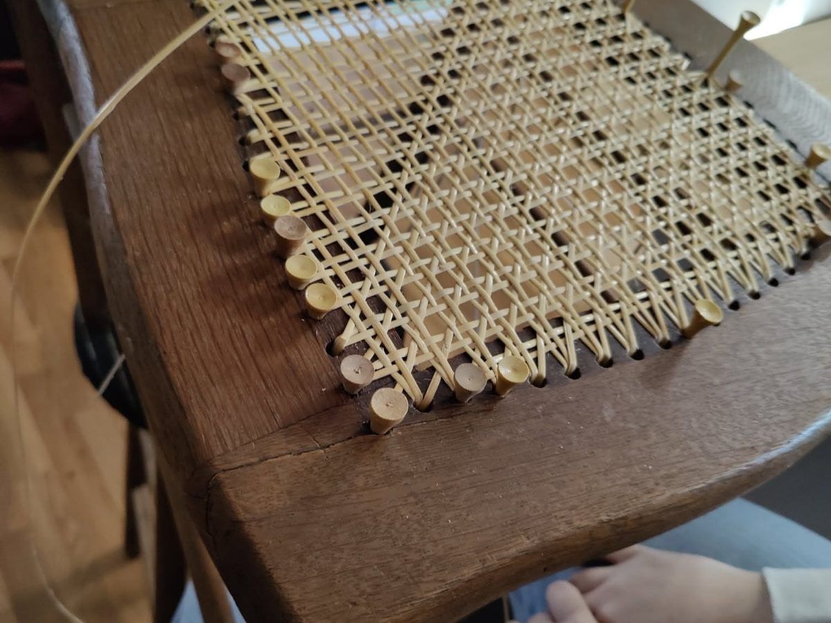 A chair seat being woven by hand in cane, the pattern half finished and loose strands still hanging from the frame.