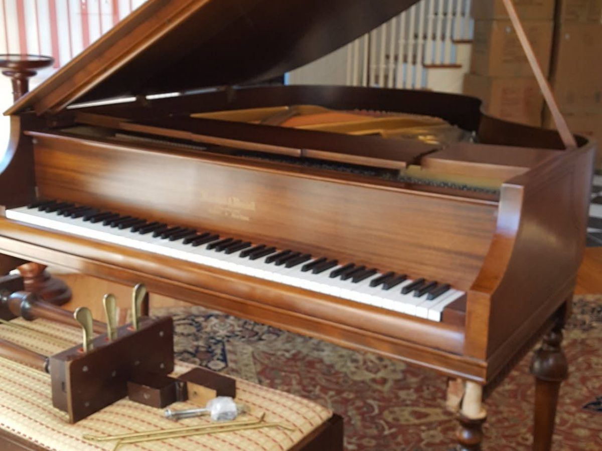 A grand piano part-way through restoration, its lid raised and the keyboard exposed, with dismantled action parts laid out on a bench in front of it.